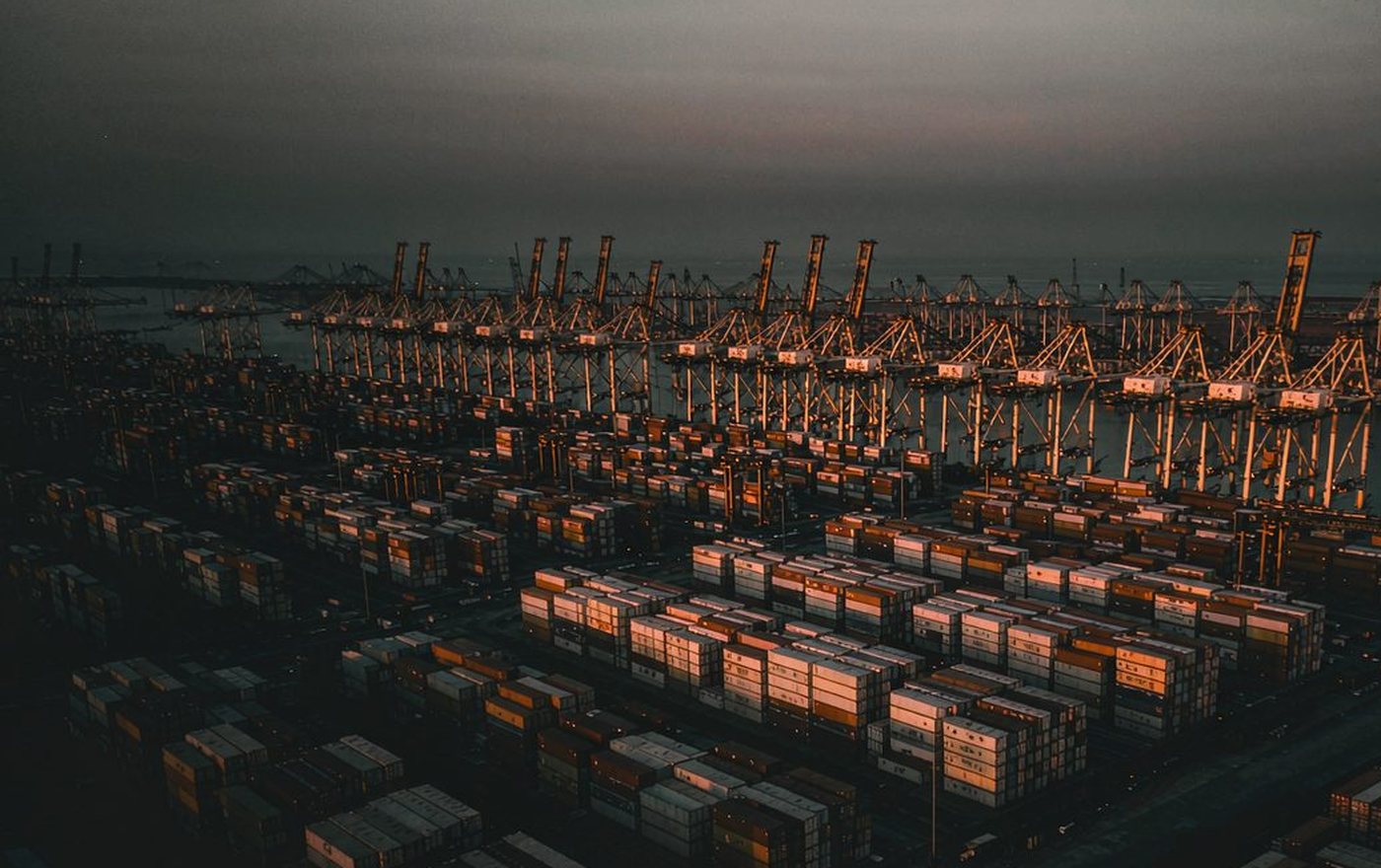 Shipping containers stacked at a container port at dusk beneath rows of gantry cranes